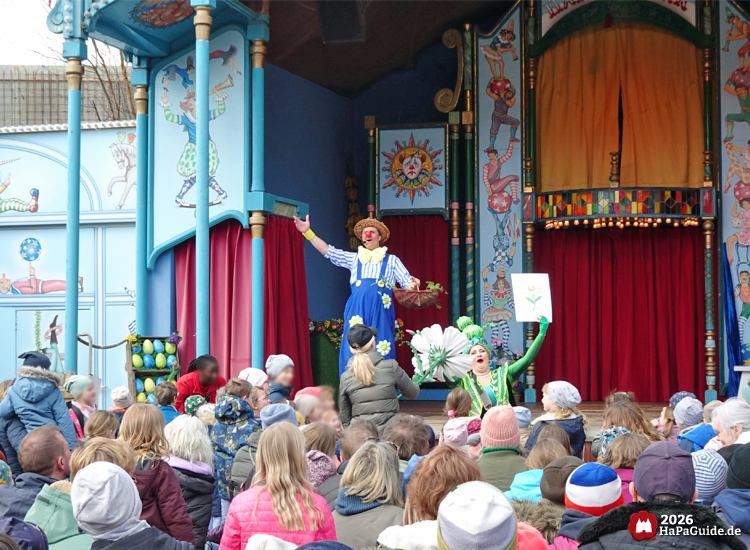 Schaubudenzauber - Die Raupe hält ein Foto einer Blume hoch während eine Frau im Publikum eine Blume in der Hand hält