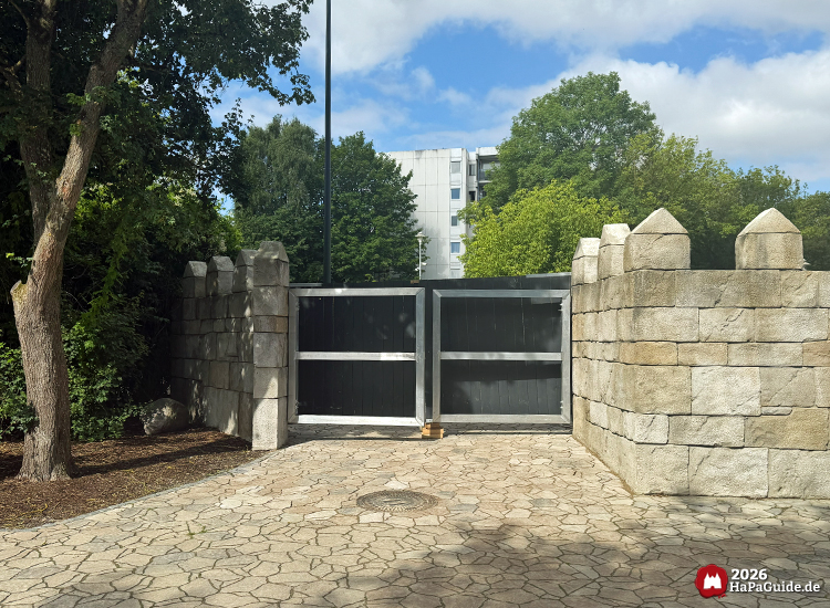 Stahlkonstruktion vor schwarzen Doppeltüren am hinteren Bereich des Palacio de Braga im Hansa-Park, flankiert von massiven Steinmauern und umgeben von Bäumen und Pflasterweg