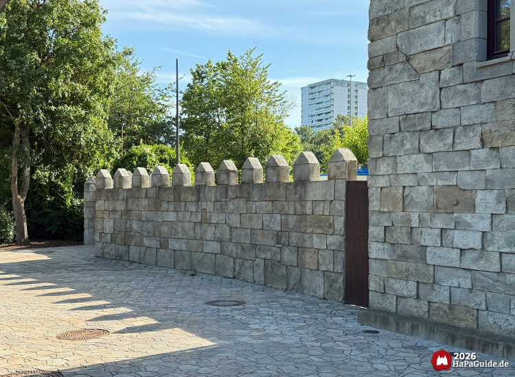 Steinmauer des Palacio de Braga im Hansa-Park mit gleichmäßig gesetzten Zinnen, dahinter grüne Bäume und Hochhaus im Hintergrund unter blauem Himmel