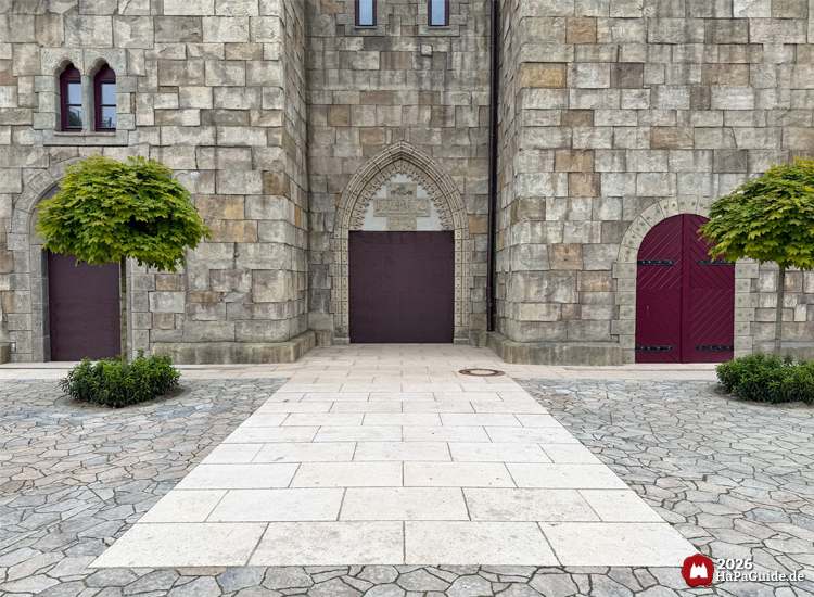 Steinerne Burgfassade des Palacio de Braga im Hansa-Park mit drei Toren, spitzbogigem Mittelportal und zwei symmetrisch gepflanzten Kugelbäumen auf gepflastertem Vorplatz