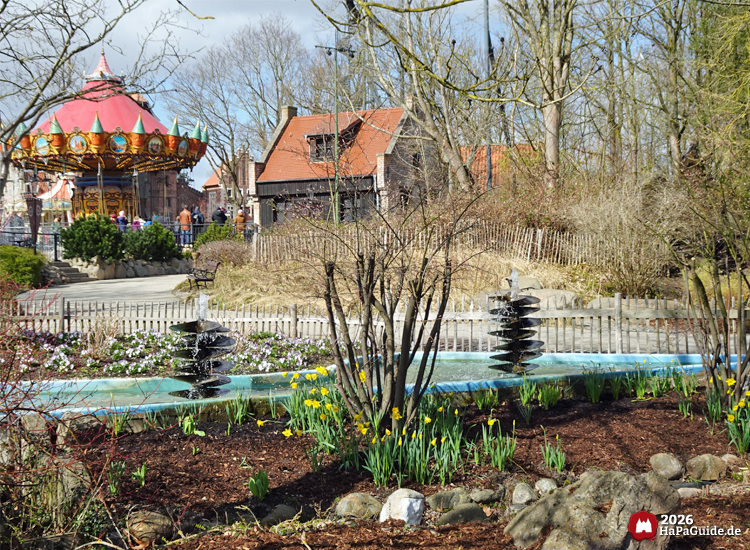 Frühlingshafte Parkszene mit Narzissen, Wasserfontänen und Hanse-Flieger vor rustikalen Gebäuden im Hansa Park