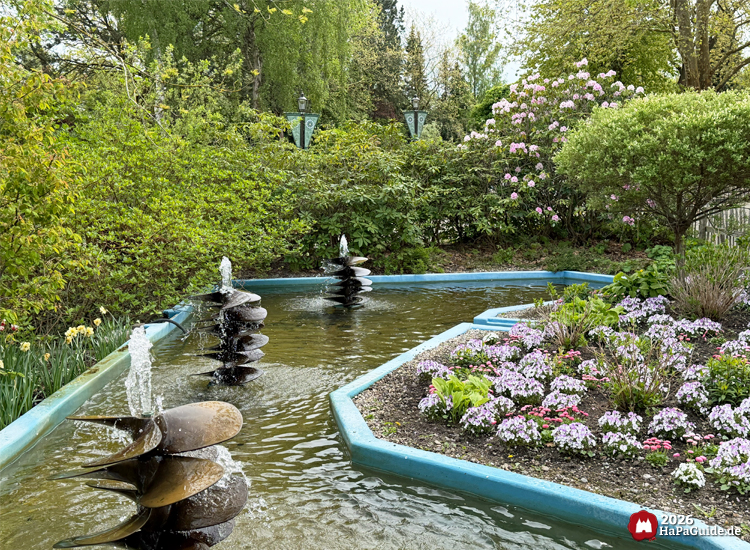 Spiralförmige Schiffschrauben in einem Teich im Hansa-Garten, umgeben von blühenden Blumenbeeten und dichtem Grün im Hansa-Park