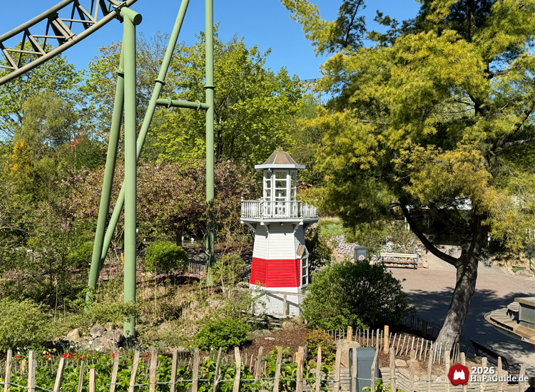 Kleiner weiß-roter Leuchtturm im Hansa-Park, umgeben von grünen Bäumen und Sträuchern, mit Teilen der Achterbahnstrecke Flucht von Novgorod im Hintergrund