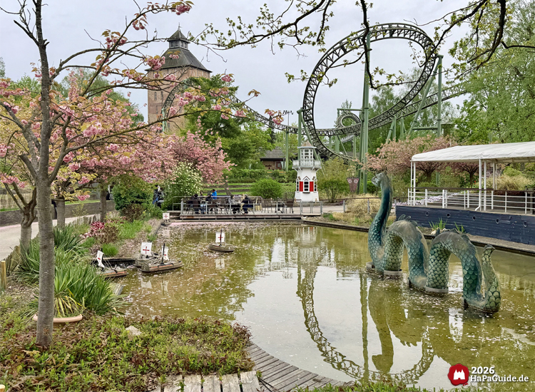 Teich im Hansa-Garten mit grüner Nessie-Skulptur und kleinen Fernlenkschiffen, umgeben von blühenden Kirschbäumen und der Achterbahn Flucht von Novgorod im Hintergrund