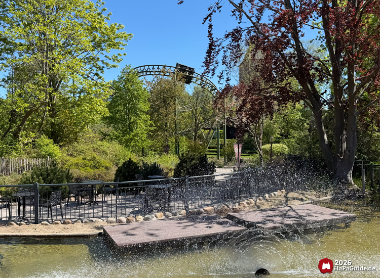 Spritzendes Wasserbecken mit Holzplattformen im Hansa-Park, umgeben von grünen Bäumen und Sträuchern, im Hintergrund die Achterbahn Flucht von Novgorod unter blauem Himmel