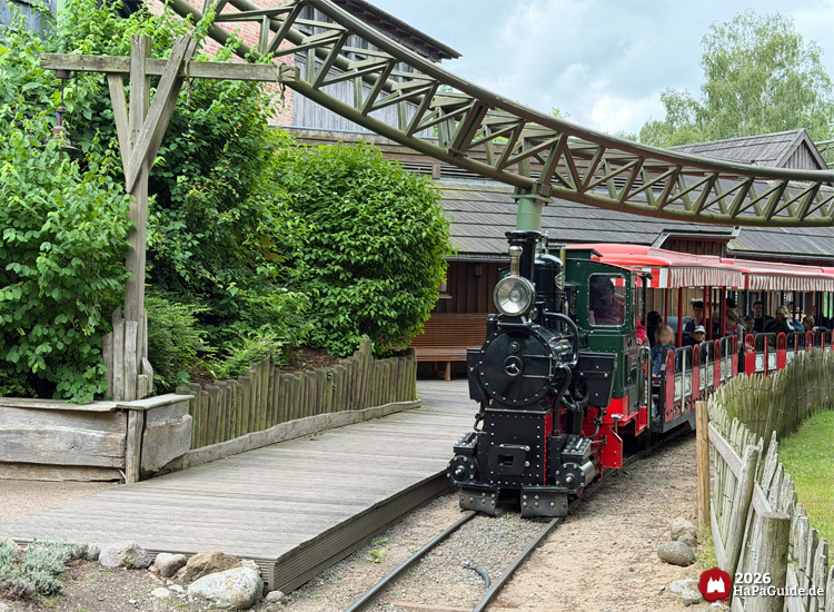 Grün-rote Dampflokomotive der Parkbahn im Hansa-Park steht unter der Achterbahn Flucht von Novgorod am Bahnhof Peterhof von Novgorod, umgeben von grünen Büschen und Holzzäunen