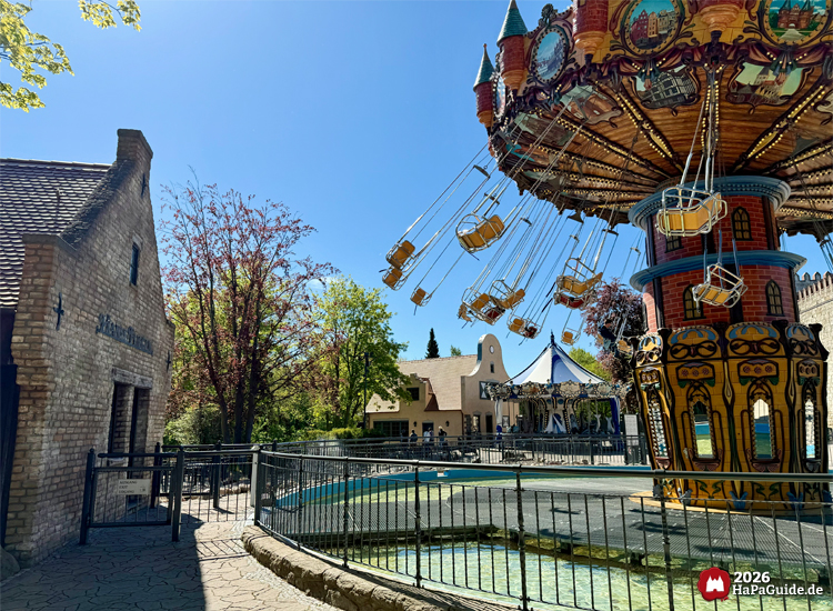 Kettenkarussell Hanse-Flieger im Hansa-Park mit reich verziertem Sockel und beigen Sitzen, daneben das Operator Gebäude und das maritime Carrousel Baltique, umgeben von Bäumen unter blauem Himmel