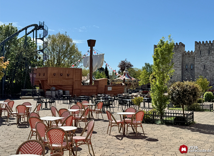 Hölzerne Hansekogge mit weißen Segeln und bunten Wimpeln in der Hanse in Europa im Hansa-Park, umgeben von Café-Tischen und Stühlen, im Hintergrund das graue Burggebäude Palacio de Braga und die Achterbahn Nessie