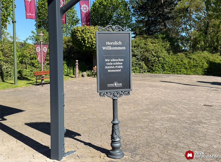 Schwarzes Willkommensschild im Hansa-Park mit der Aufschrift 'Herzlich Willkommen – Wir wünschen viele schöne HANSA-PARK-Momente!', umgeben von grünen Bäumen und roten Bannern