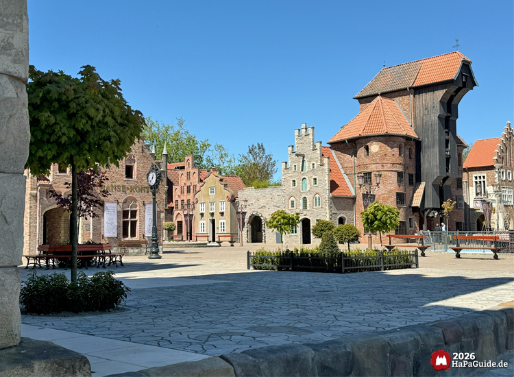 Blick durch die Hanse in Europa im Hansa-Park mit historischen Backstein- und Steinfassaden, Türmen und Giebeln, im Vordergrund gepflasterter Platz und gepflegte Grünflächen unter blauem Himmel