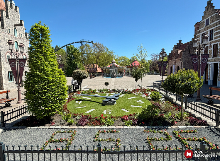 Blumenuhr hinter dem Eingang zum Hansa-Park mit grüner Rasenfläche und weißen Zahlen, umgeben von bunten Blumenbeeten und historischen Gebäuden, im Hintergrund das Hanse-Karussell