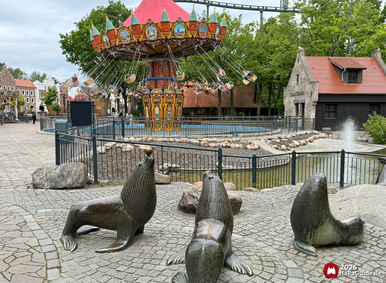 Hanse-Flieger im Hansa-Park mit rotem Dach und bunten Ornamenten, davor drei bronzene Seelöwenfiguren auf Kopfsteinpflaster neben einem kleinen Teich mit Springbrunnen