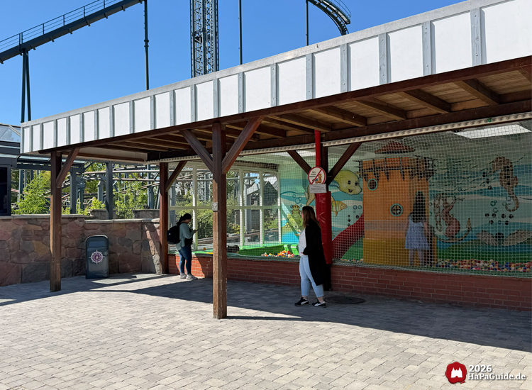 Überdachter Spielbereich im Hansa-Park mit Unterwasserwand, gelbem Turm und roter Rutsche im Ballbecken, beobachtet von Besuchenden bei sonnigem Wetter