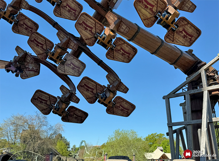 Attraktion Kärnapulten im Hansa-Park mit hölzernen Flugapparaten, die sich in der Luft drehen, vor blauem Himmel und grünen Bäumen