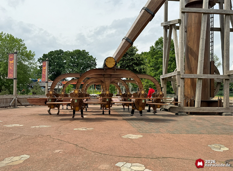 Basis des Rundfahrgeschäfts Kärnapulten im Hansa-Park mit hölzernen Fluggeräten und großem Katapultfuß, umgeben von roten Bannern und gepflastertem Platz