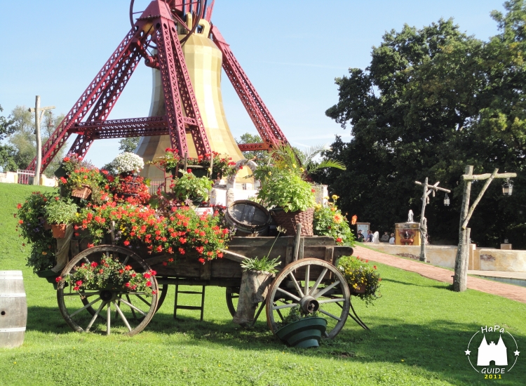 Zahlreiche Blumen auf einer Holzkutsche vor der Glocke