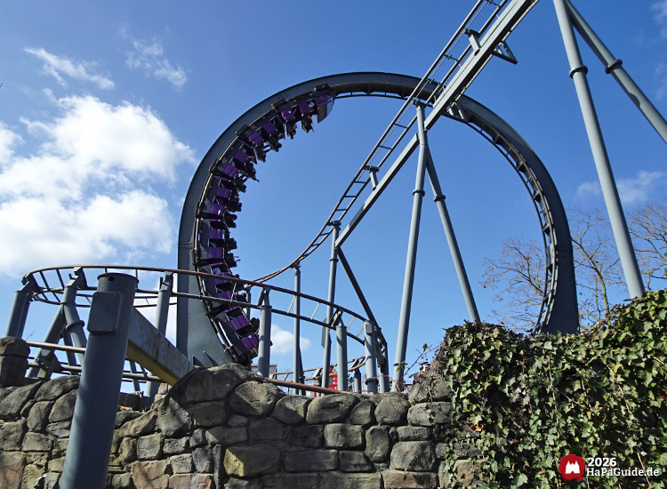 Stahlachterbahn Nessie im Hansa-Park mit dunkelgrauem Looping und lilafarbenem Zug vor blauem Himmel und Steinmauer mit Efeu