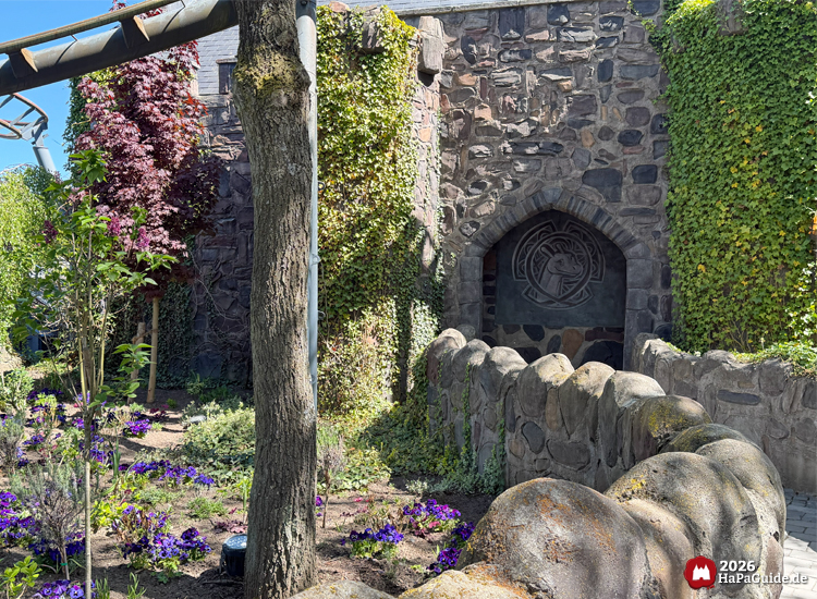 Steinmauer mit Torbogen und Reliefmotiv Nessies am Eingang zum Eilean Donan Castle im Hansa-Park, umgeben von Efeu, Blumen und Bäumen