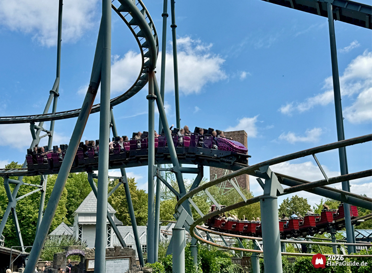 Zug der Loopingbahn Nessie im Hansa-Park fährt aus der geschwungenen Helix mit abwärts, im Hintergrund grüne Bäume, der Royal Scotsman und Gebäude des Themenbereichs