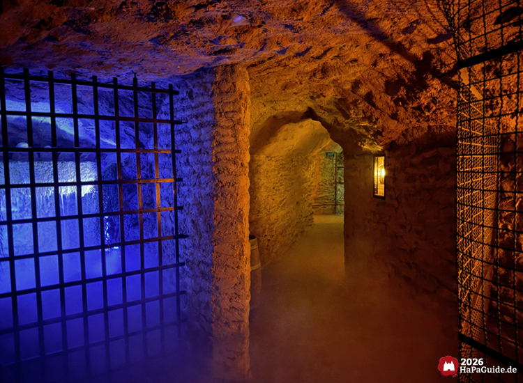 Steiniger Tunnel der Bärenhöhle des Hansa-Parks mit blauer und orangefarbener Beleuchtung, Nebel und Gittertoren, vermittelt eine geheimnisvolle Stimmung