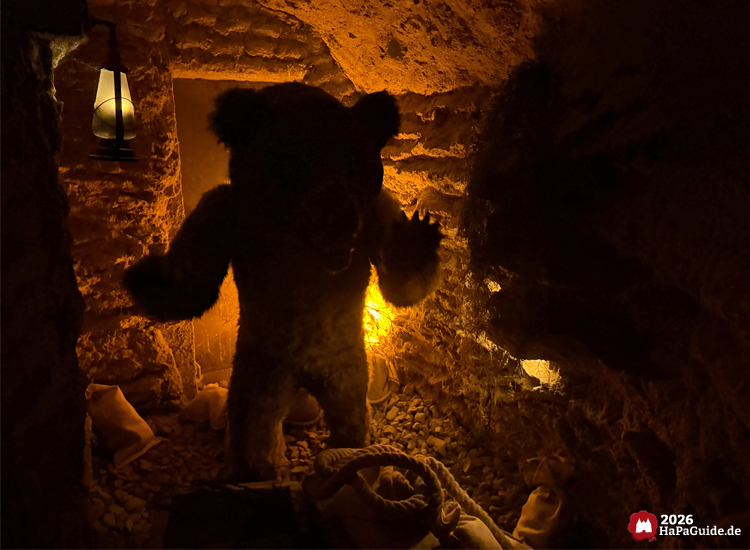 Bärenfigur in der Bärenhöhle des Hansa-Parks steht in einem dunklen Raum mit Laternenlicht und steinernen Wänden, umgeben von Seilen und Säcken, wirkt lebensecht und eindrucksvoll