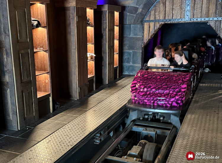 Zug der Achterbahn Nessie im Hansa-Park fährt mit neuer Frontfolierung aus dem dunklen Tunnel in die Station mit beleuchteten Holzschränken und Steinwänden, Wagen mit violettem Schuppenmuster und Fahrgästen