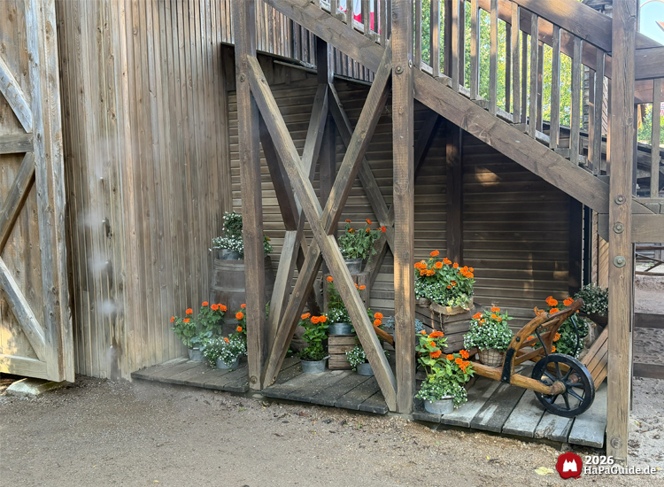 Fort im Westernbereich New Lübeck des Hansa-Parks mit Blumendekoration aus orangefarbenen Ringelblumen und weißen Blüten in Holzfässern und Kisten, daneben ein rustikaler Handkarren und eine Wand aus Wassernebel