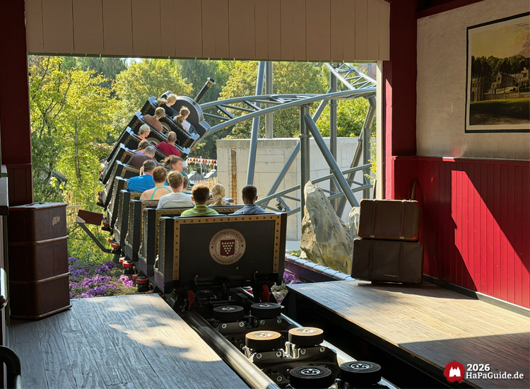 Cornwall Coaster mit Wappen beim Ausfahren aus der Station, umgeben von Felsen und Bäumen im Hansa-Park