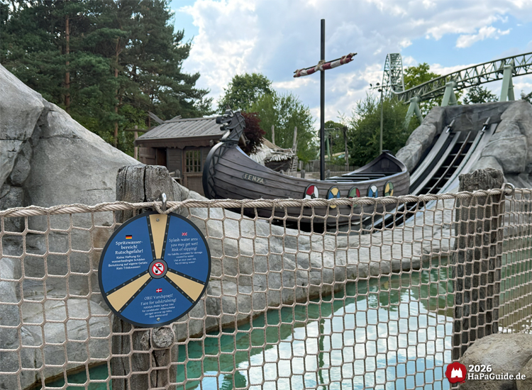 Wikinger-Boot Fenja von Einars Fjordfahrt im Wasserbecken mit Felskulisse, Hinweisschild zum Spritzwasserbereich und Achterbahn im Hintergrund im Hansa-Park