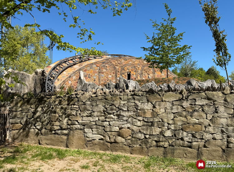 Steinmauer mit gezacktem Abschluss und dahinterliegender Rundbogenstruktur des Cornwall Coaster 2026 im Themenbereich Bezauberndes Britannien des Hansa-Parks – sonniger Frühlingstag mit blühenden Bäumen