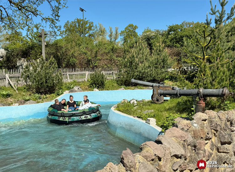 Besuchende in einem Rundboot des Wasser-Raftings Störtebekers Kaperfahrt im Hansa-Park, umgeben von Felsen, grüner Vegetation und thematisierter Kanone bei sonnigem Himmel