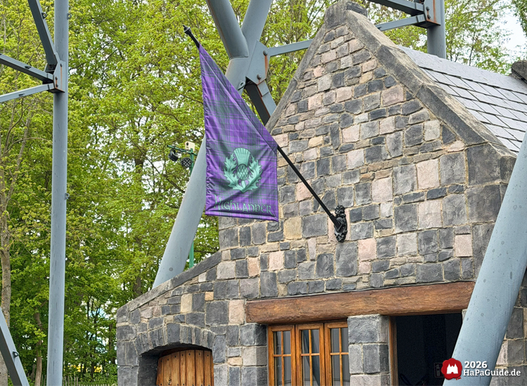 Steingebäude der Highlander-Attraktion im Hansa-Park mit lila karierter Flagge und grünem Distelmotiv, umgeben von Metallstützen und Bäumen