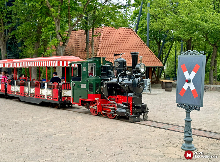 Grün-rote Parkbahn Hansa-Park Express mit Dampflokomotive im Hansa-Park fährt vor einem Bahnübergangsschild mit rotem Kreuz, dahinter Bäume und der Kiosk am Hanse-Flieger