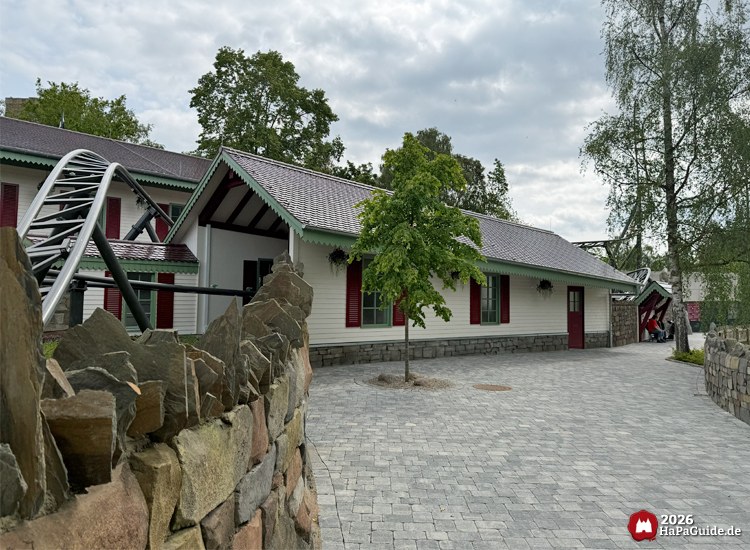 Helle Gebäude im Cornwall Coaster Bereich des Hansa-Park mit roten Fensterläden und grünen Dachkanten, gepflasterter Weg und graue Steinmauer im Vordergrund unter bewölktem Himmel