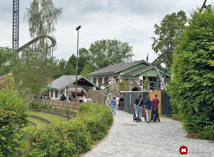 Fußweg im Hansa-Park führt entlang der Achterbahn Cornwall Coaster mit weißen Gebäuden, grünen Dachkanten und roten Fensterläden, umgeben von Bäumen und Sträuchern unter grauem Himmel