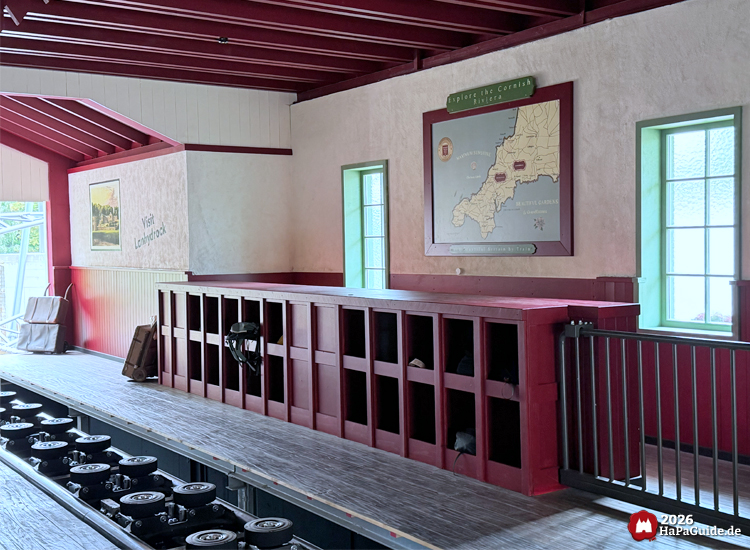 Innenbereich der Achterbahnstation Cornwall Coaster im Hansa-Park mit roten Gepäckablagefächern, grünen Fenstern und einer Karte der 'Cornish Riviera' an der Wand, daneben ein Poster 'Visit Lanhydrock'