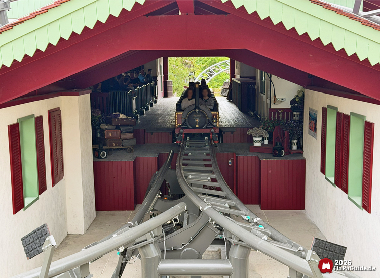 Achterbahn Cornwall Coaster im Hansa-Park verlässt die Station mit grauer Schienenkonstruktion, rotem Dach und weißen Wänden mit grünen Fensterrahmen, im Hintergrund wartende Fahrgäste