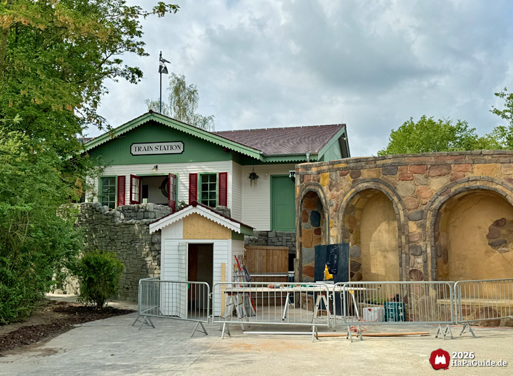 Cornwall Coaster Bahnhofsgebäude mit der Aufschrift 'Train Station' im Hansa-Park mit grünen Dachkanten und roten Fensterläden, daneben eine Steinmauer und Baustellenabsperrungen vor einem kleinen weißen Häuschen