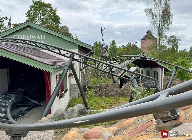 Achterbahn Cornwall Coaster im Hansa-Park mit grauer Stahlstrecke, die durch weiße Gebäude mit grünen Dachkanten und roten Fensterläden führt, im Hintergrund der Saviour-Tower und grüne Bäume