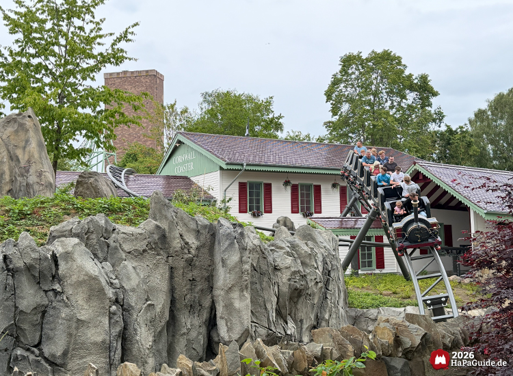 Achterbahn Cornwall Coaster im Hansa-Park fährt mit schwarzer Lokomotive und Fahrgästen über graue Felsen, dahinter das Stationsgebäude mit grünen Dachkanten und roten Fensterläden unter bewölktem Himmel