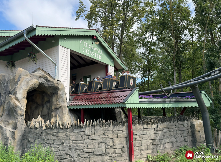 Station der Cornwall Coaster im Hansa-Park mit grünem Giebel, rotem Dach und Felsen mit Höhleneingang, Zug mit Fahrgästen bereit zur Abfahrt unter blauem Himmel