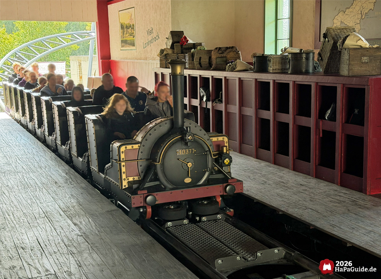 Zug der Cornwall Coaster Achterbahn im Hansa-Park fährt durch die Station mit roten Holzregalen und Reisekoffern, Fahrgäste sitzen in schwarzen Wagen mit goldenen Verzierungen