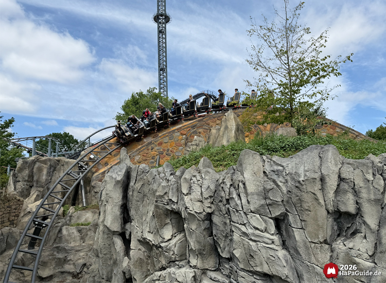Zug der Cornwall Coaster Achterbahn im Hansa-Park fährt über eine steinige Hügelkonstruktion mit Felsen und grüner Vegetation, im Hintergrund der Highlander-Turm