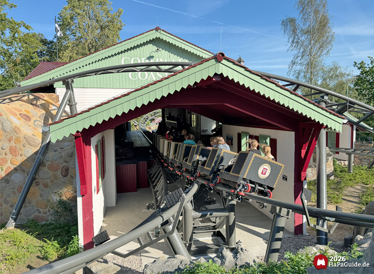 Zug der Cornwall Coaster Achterbahn im Hansa-Park fährt in die Station mit grünem Dach und rotem Rahmen, schwarze Schienen führen durch die Landschaft unter blauem Himmel