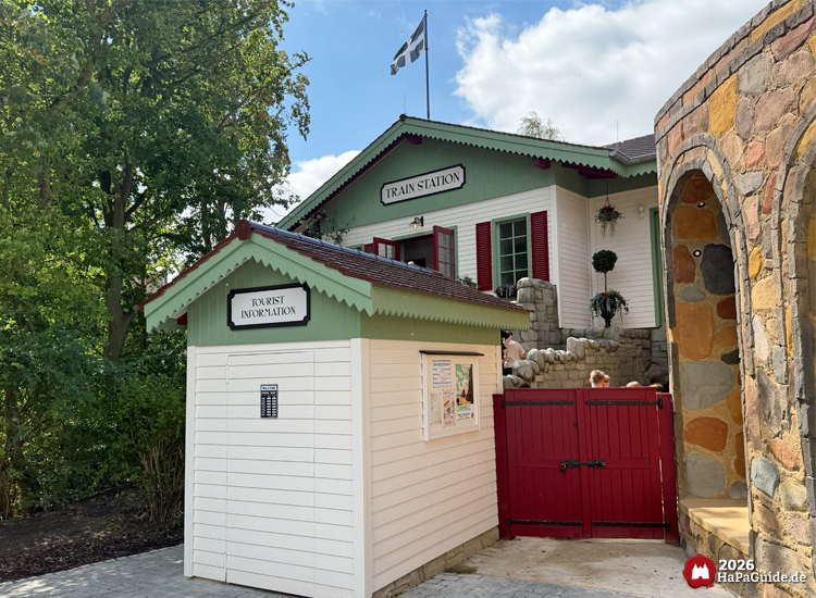 Gebäude der Cornwall Coaster Station im Hansa-Park mit Schildern 'Train Station' und 'Tourist Information', weißen Wänden, roten Fensterläden und grüner Dachkante