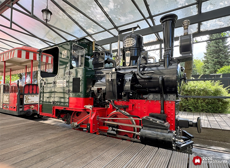 Schwarz- rote Dampflokomotive der Parkbahn im Hansa-Park steht unter Glasdach, dahinter roter und weißer Waggon mit goldenen Verzierungen, Hauptbahnhof Hansa-Garten mit Holzplattform