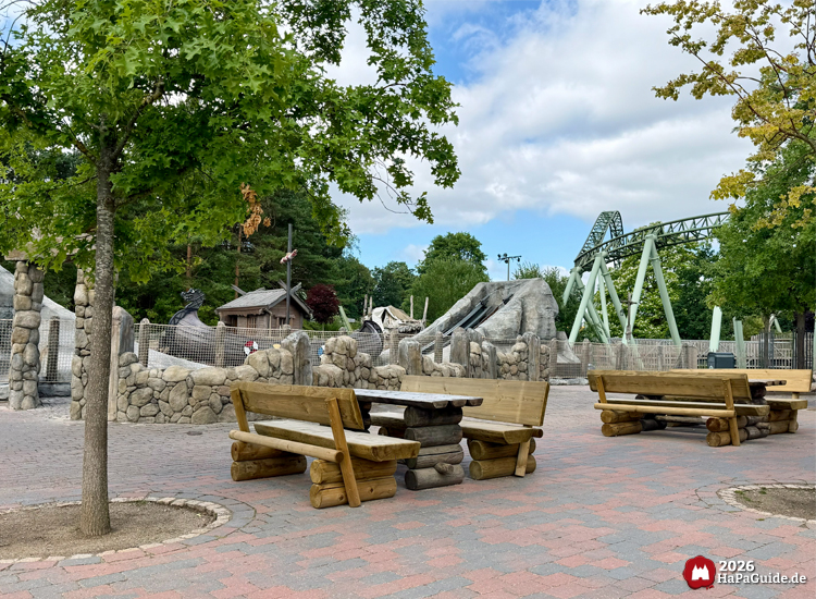 Sitzbereich mit Holzbänken und Tischen im Wikingerland des Hansa-Park, umgeben von Bäumen und Blick auf Wikinger-Taufe mit Felsen und Einars Fjordfahrt im Hintergrund