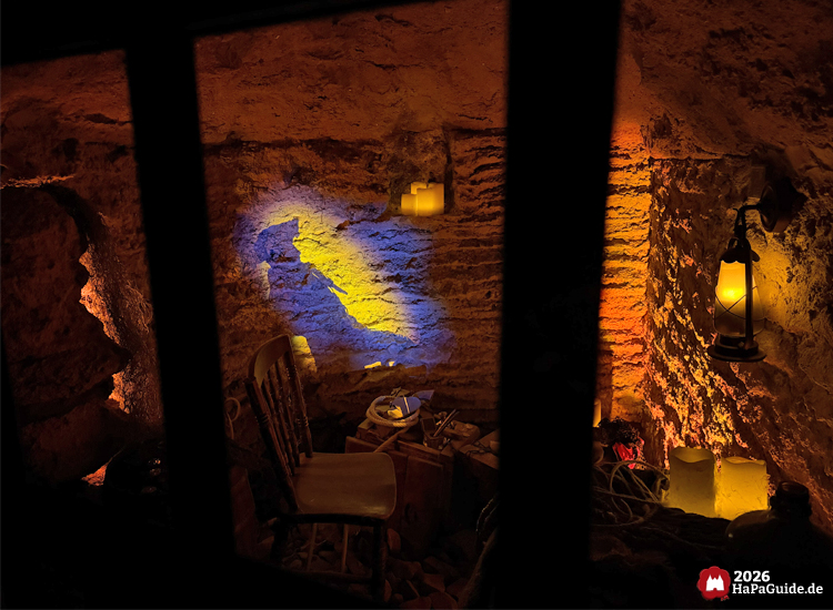Unterirdischer Raum in der Bärenhöhle des Hansa-Parks mit Holzstuhl, Kerzen und Laternen, warmes orangefarbenes Licht und Projektion eines Seemanns an der Steinwand erzeugen eine mystische Atmosphäre