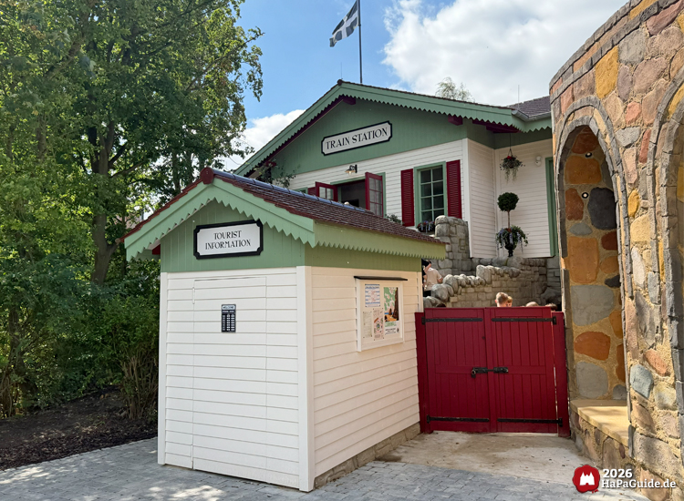 Tourist Information-Hütte vor dem Gebäude Train Station im Cornwall Coaster-Bereich des Hansa-Parks, weiß-grüne Fassade mit roten Fensterläden und Steinmauer, sonniger Tag
