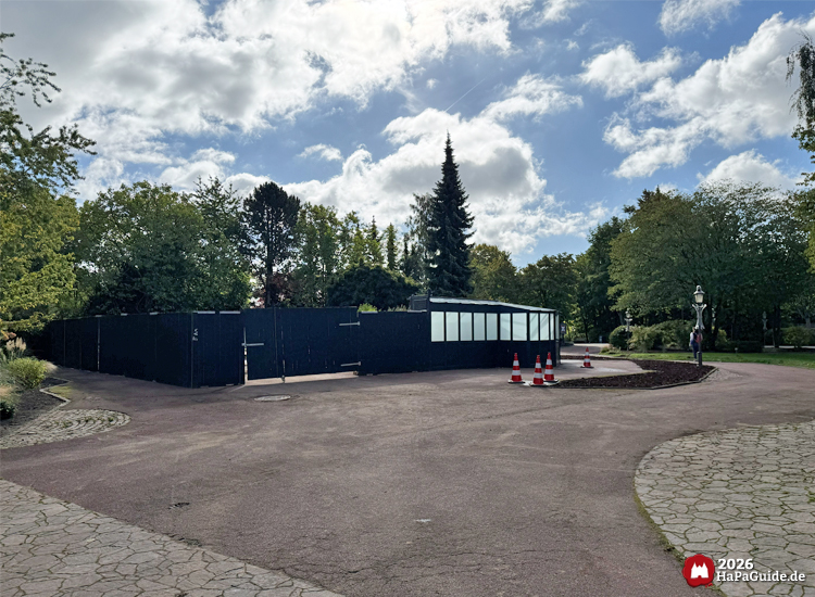 Baustellenbereich des Buddenbrook-Turms im Hansa-Park mit schwarzer Absperrung und ehemaligem Holsteinturm-Gebäude, umgeben von Bäumen und Lampen, sonniger Himmel mit Wolken über dem Parkgelände